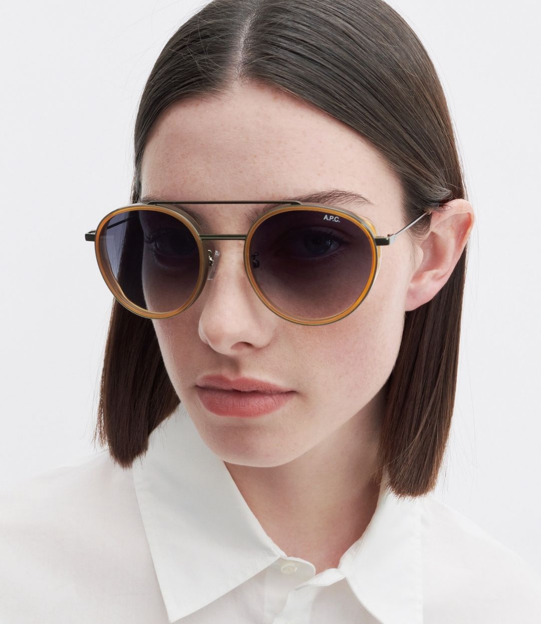 Close-up of a woman with dark hair and fair skin wearing round sunglasses with a tortoiseshell rim and a white collared shirt.