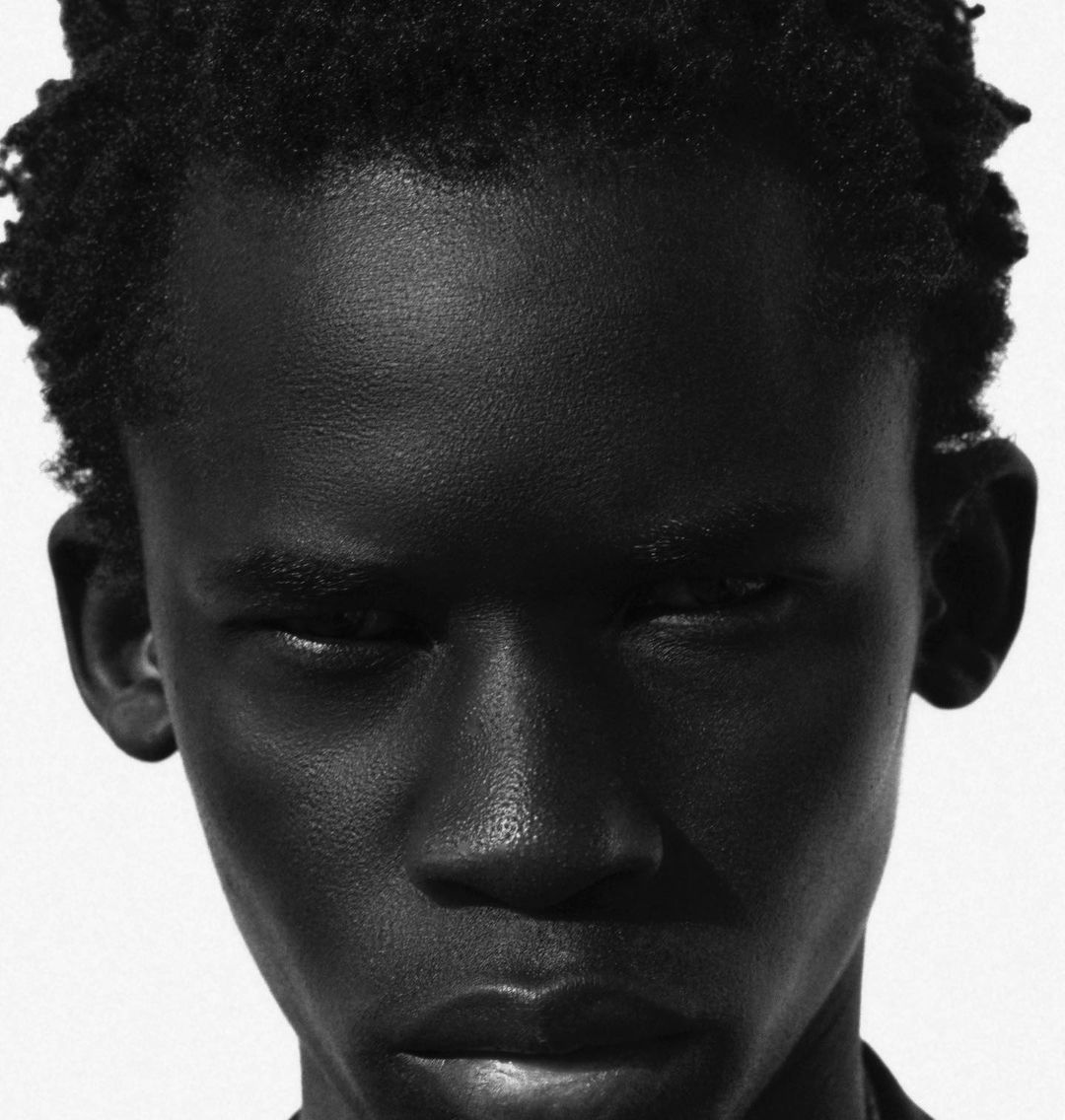 Close-up portrait of a young Black man with short curly hair in high contrast black and white lighting against a stark white background.