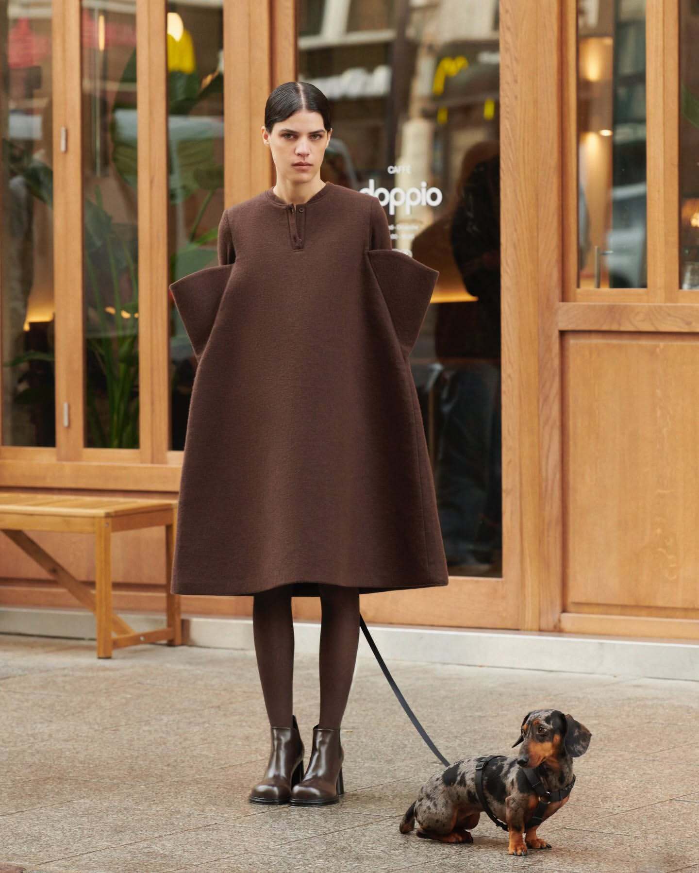 Adult female in a brown dress and boots walking a dapple dachshund in an urban setting in front of a cafe with wood accents.