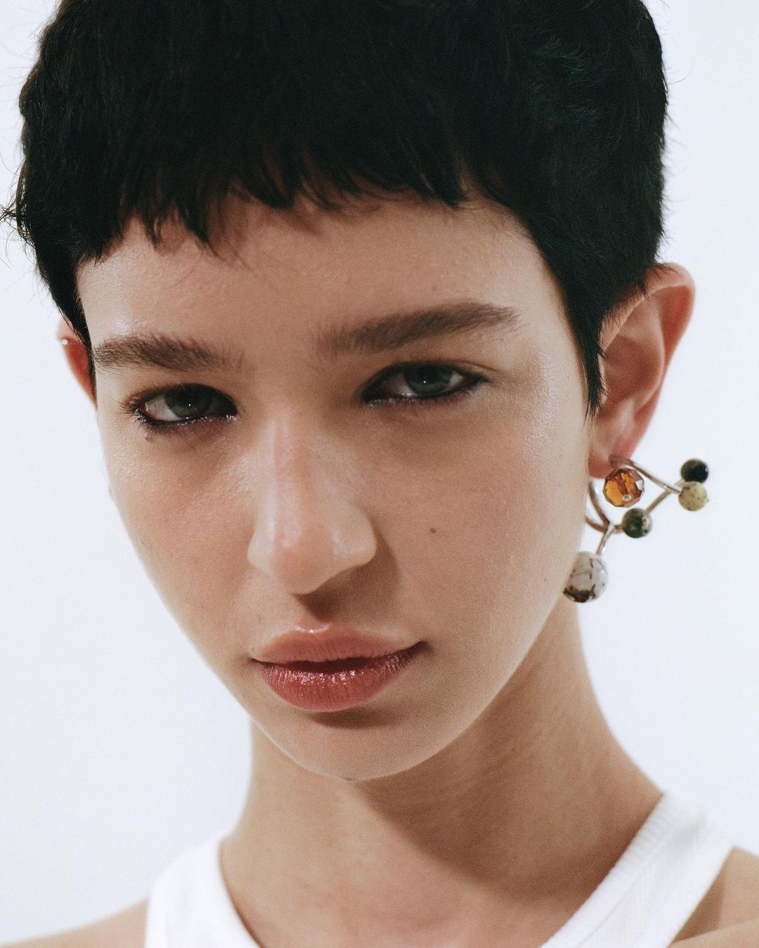 Close-up shot of a woman with short dark hair and fair skin, wearing a white top and statement earrings, set against a clean white background.