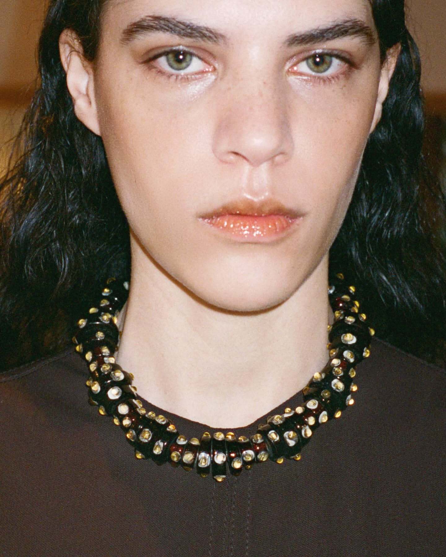 Close-up of a brunette woman with fair skin wearing a beaded statement necklace against a neutral background in eye-level view.