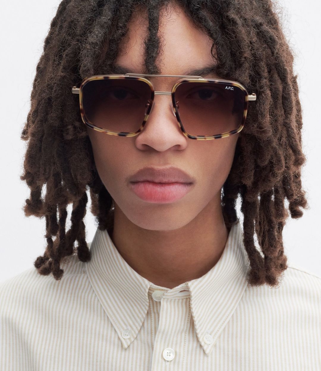 Close-up portrait of a young adult wearing tortoiseshell sunglasses and a striped button-down shirt against a neutral background.