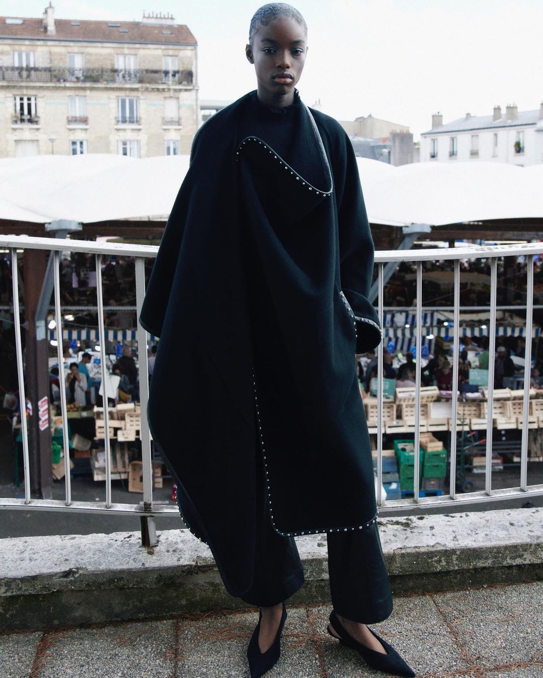 Runway shot of a black female model wearing a large, black avant-garde coat with silver embellishments, posing outdoors in front of a crowd.