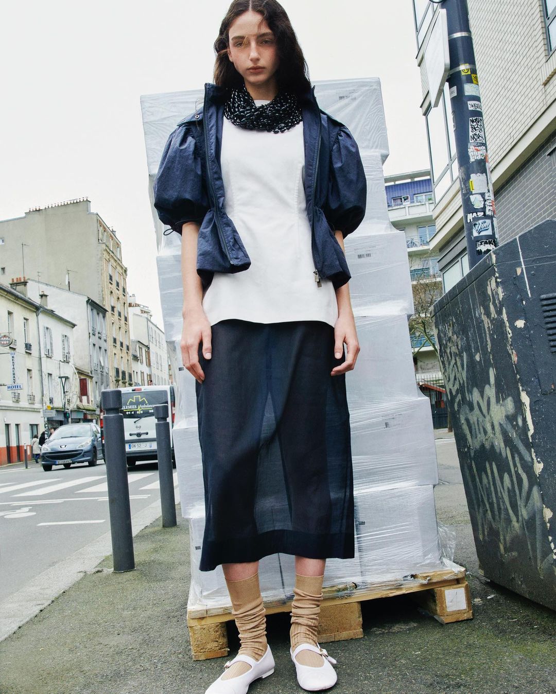 Fashionable young woman posing on urban street wearing a navy blue jacket, white top, and long skirt, accessorized with white shoes and tan socks.