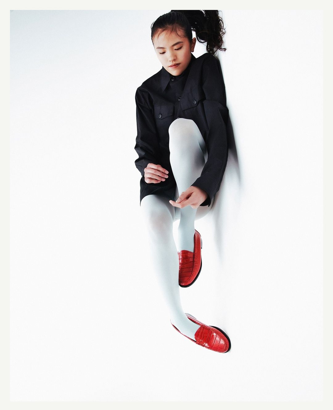 A young Asian woman with long black hair poses in a black shirt dress, white tights, and red leather shoes against a white backdrop.