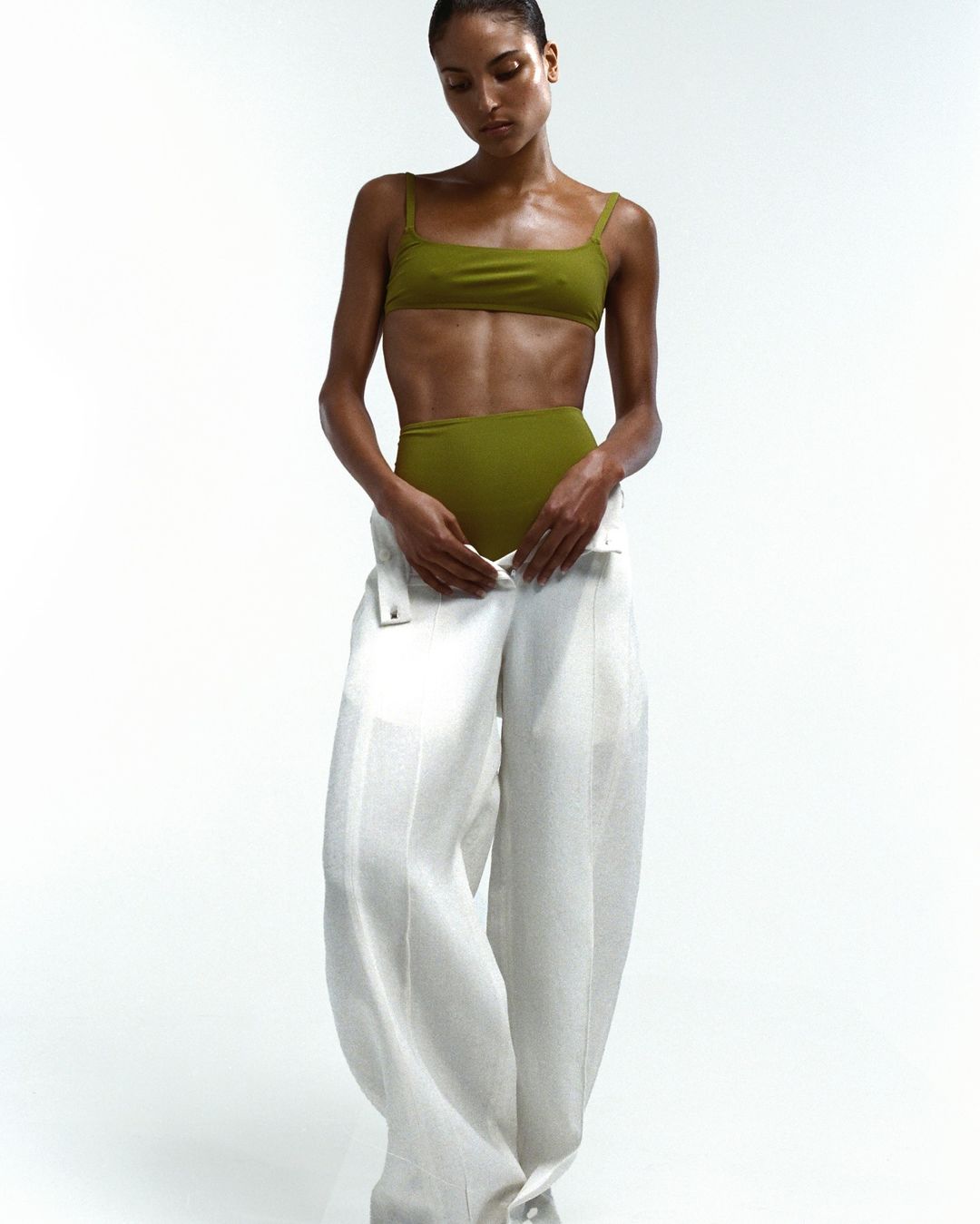 A female fashion model poses in a studio wearing a green top and large, white linen pants against a white background.