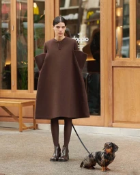 Model in oversized structured brown dress standing on a sidewalk with a dachshund on a leash outside a wooden storefront.