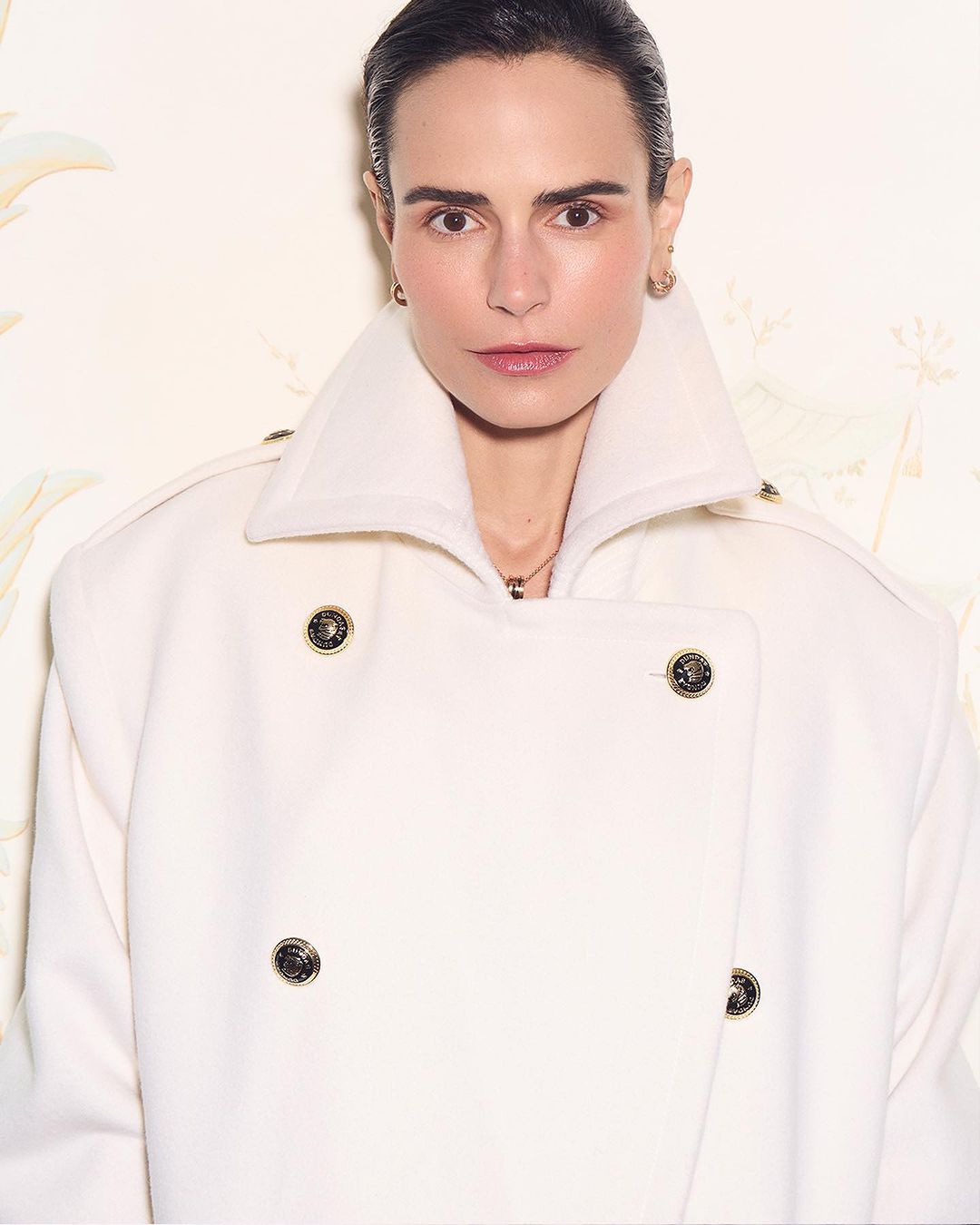 Close-up of a woman in a white double-breasted coat with metallic buttons, standing indoors against a light patterned wall.