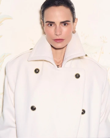 Woman wearing a heavy white double-breasted coat with large gold buttons against a pale patterned background
