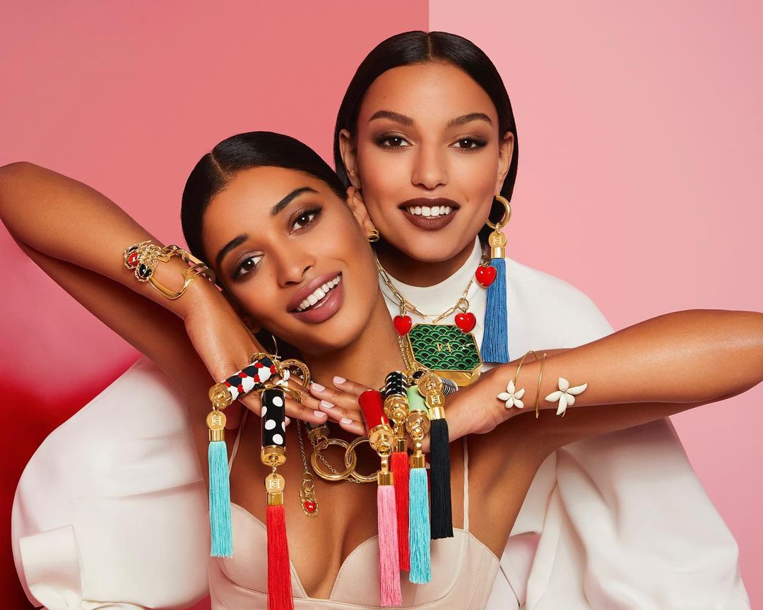 Two women pose in front of a split pink and red background, wearing ornate gold jewelry with colorful tassel and stone accents.