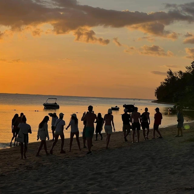 Sunset beach scene with a group of people silhouetted along the shoreline, boats visible on the water and vibrant orange and yellow sky.