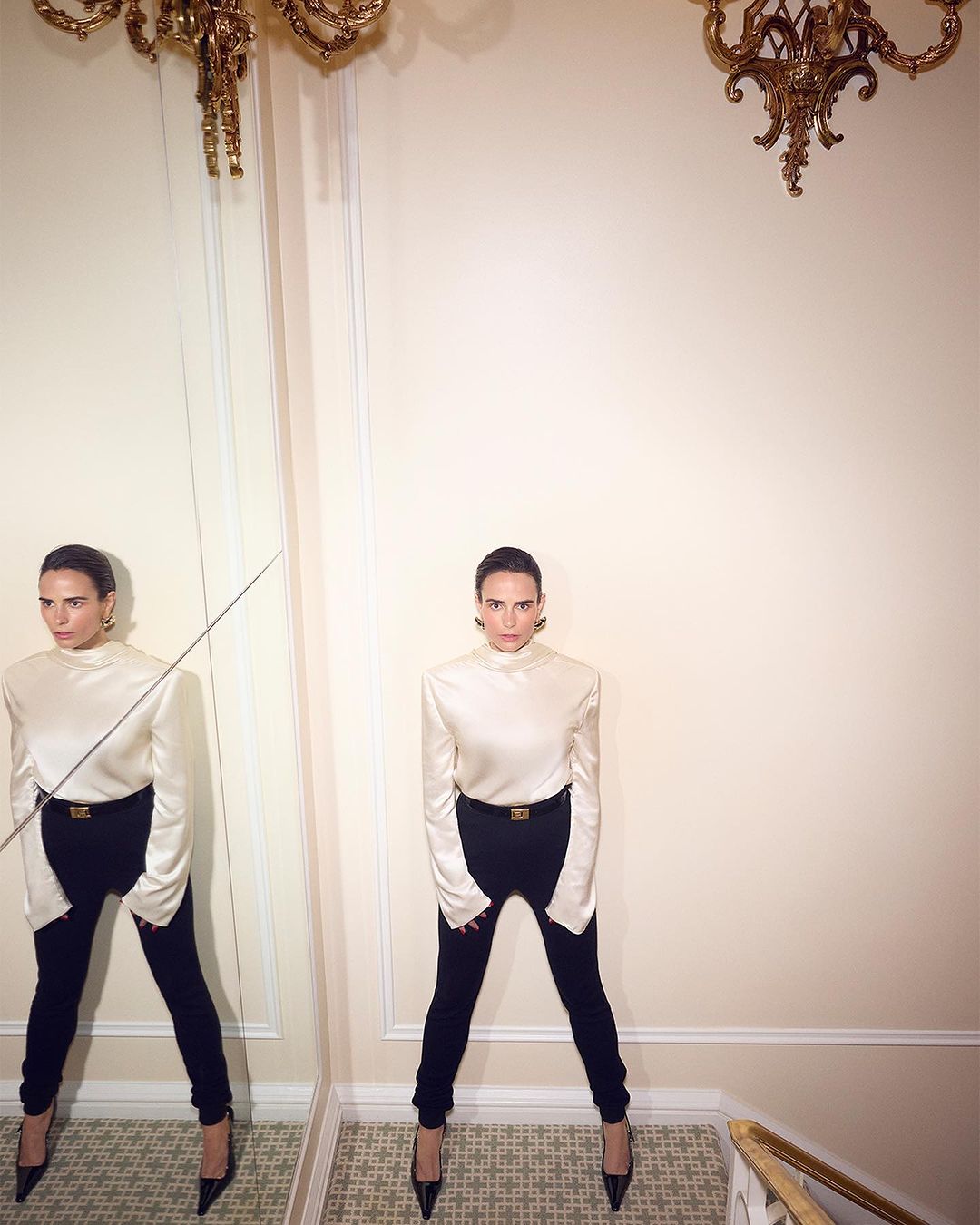 Woman posing in a cream top and black pants stands near a mirror in an elegant room with a decorative chandelier. Fashion shot.
