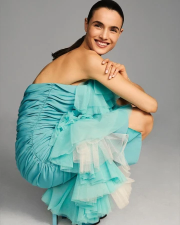 Smiling woman crouching in a bright turquoise strapless ruffled dress against a grey studio background.