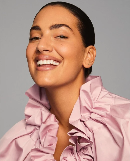 Headshot of a smiling adult woman with a pink ruffled shirt against a grey background in a studio with diffused lighting. 