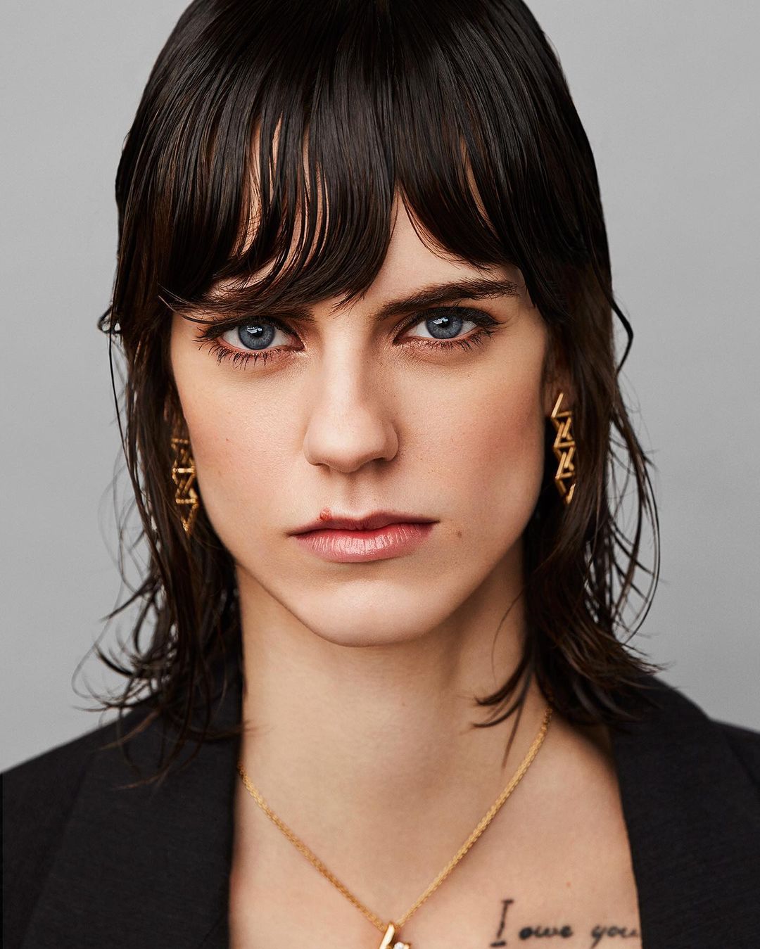 Close-up portrait of a woman with dark hair, light eyes, wearing a black blazer, gold earrings and necklace, against a gray background.