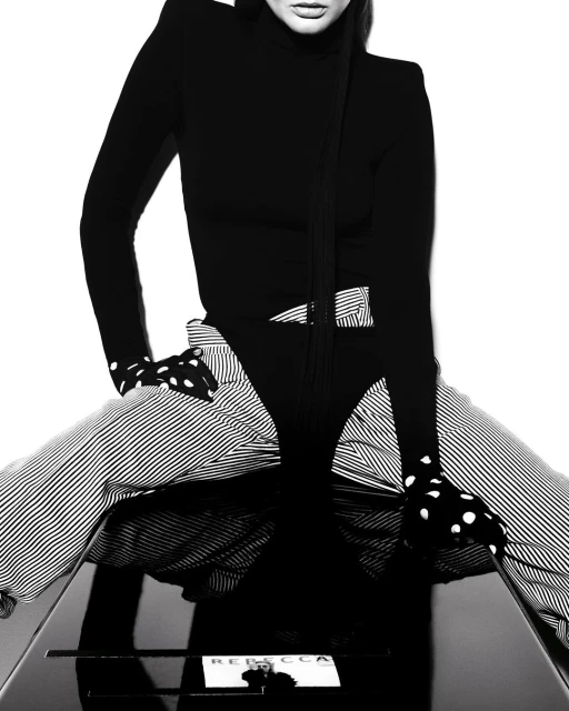 A model in black bodysuit and striped pants sits on a glossy table with a 'Rebecca' card in a black and white fashion photograph.