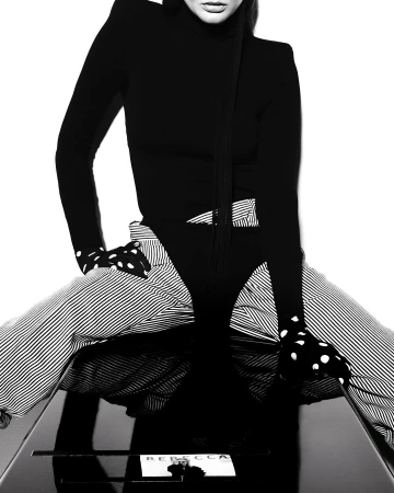 A model in black bodysuit and striped pants sits on a glossy table with a 'Rebecca' card in a black and white fashion photograph.