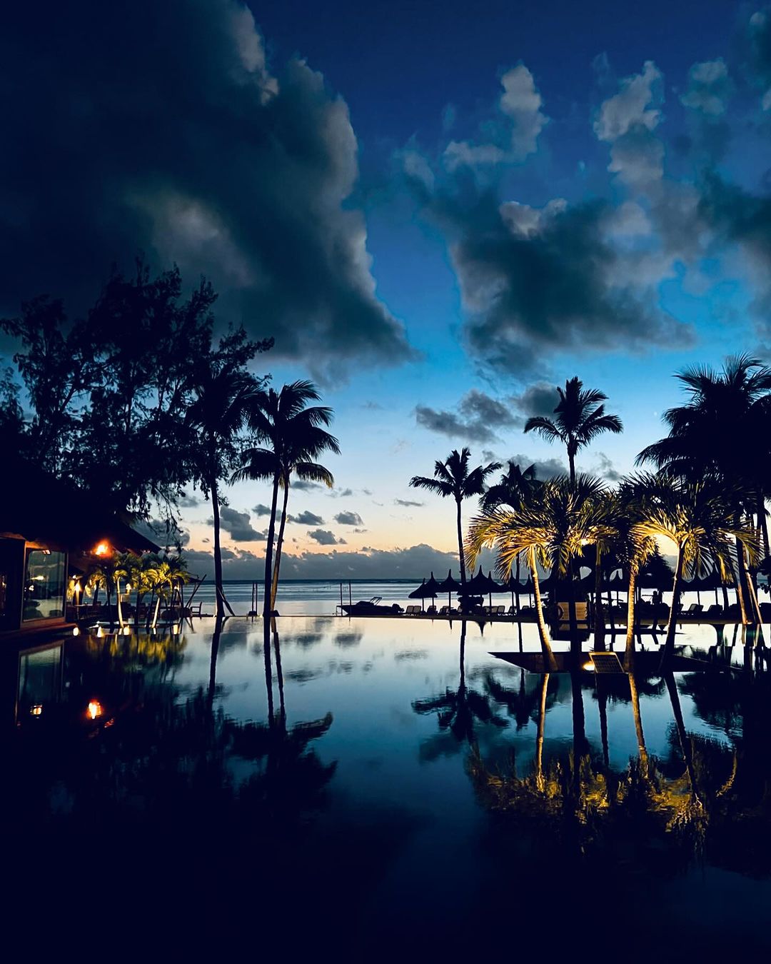 Tropical dusk scene shows calm water reflecting silhouetted palm trees, hotel structure, and a blue and cloudy sky, evoking relaxation.