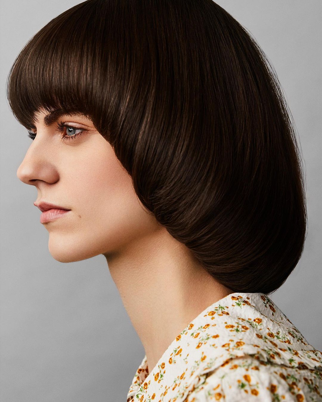 Profile portrait of an adult woman with brown blunt bob hairstyle and floral top against a gray background, focusing on her hairstyle.