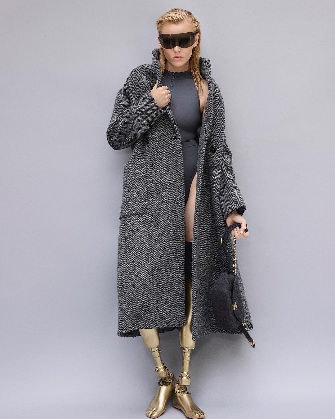 Fashion photo of blonde woman wearing a grey coat, futuristic sunglasses, and grey bodysuit with golden prosthetic legs against grey backdrop.