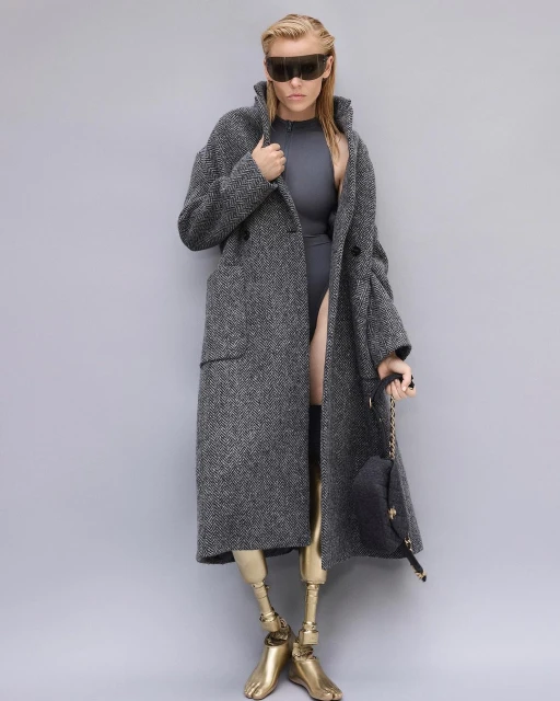 Fashion photo of blonde woman wearing a grey coat, futuristic sunglasses, and grey bodysuit with golden prosthetic legs against grey backdrop.