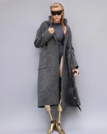 Fashion photo of blonde woman wearing a grey coat, futuristic sunglasses, and grey bodysuit with golden prosthetic legs against grey backdrop.