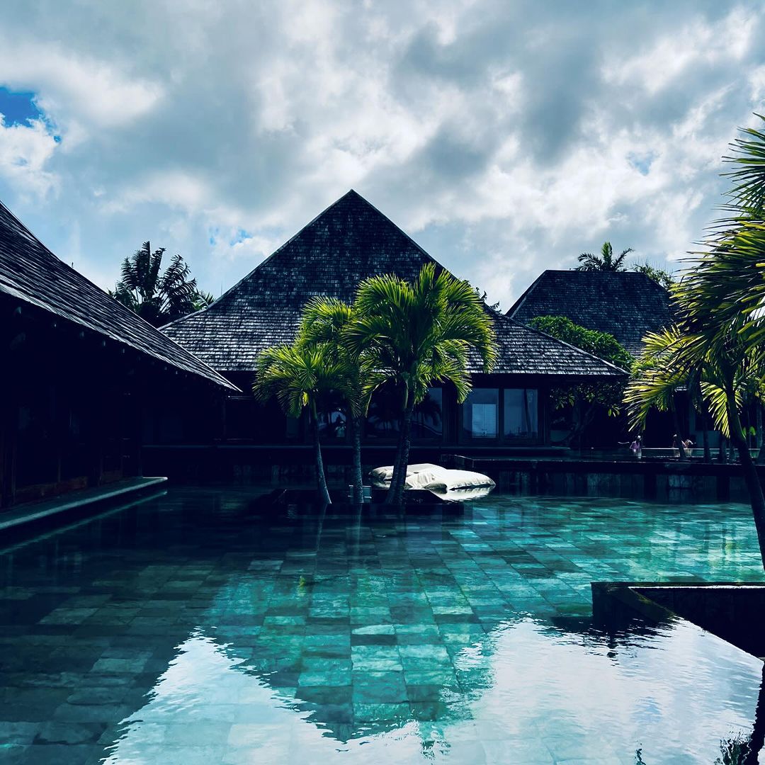 Exterior view of a tranquil pool reflecting a tropical resort with palm trees and dark wood buildings under a cloudy sky.