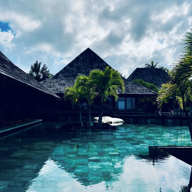 Exterior view of a tranquil pool reflecting a tropical resort with palm trees and dark wood buildings under a cloudy sky.