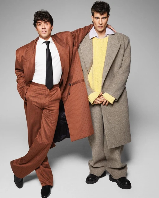 Two men in oversized coats and suits pose in a studio setting. One wears a brown pinstripe suit, the other a tan coat and black boots.