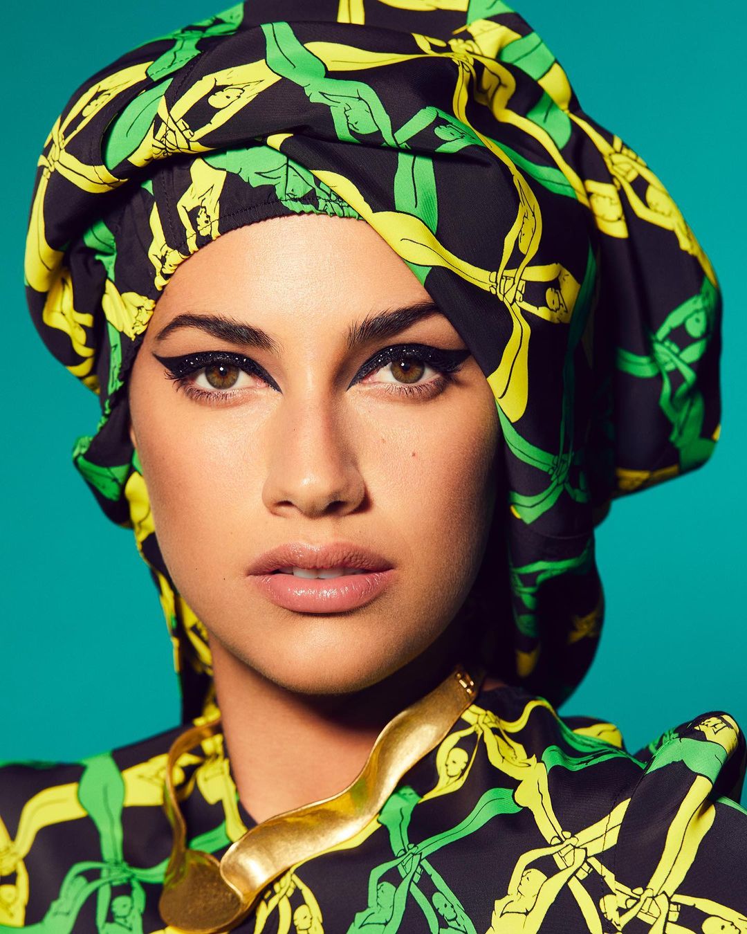 Close-up portrait of a woman with dramatic makeup, wearing a patterned headscarf and gold necklace, against a turquoise background.