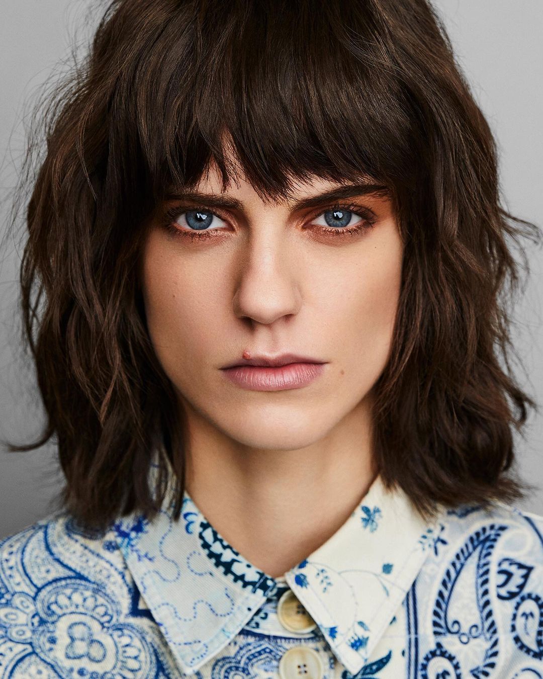 Portrait of a young adult female with brown wavy hair and blue eyes, wearing a patterned blue and white shirt, against a grey background.