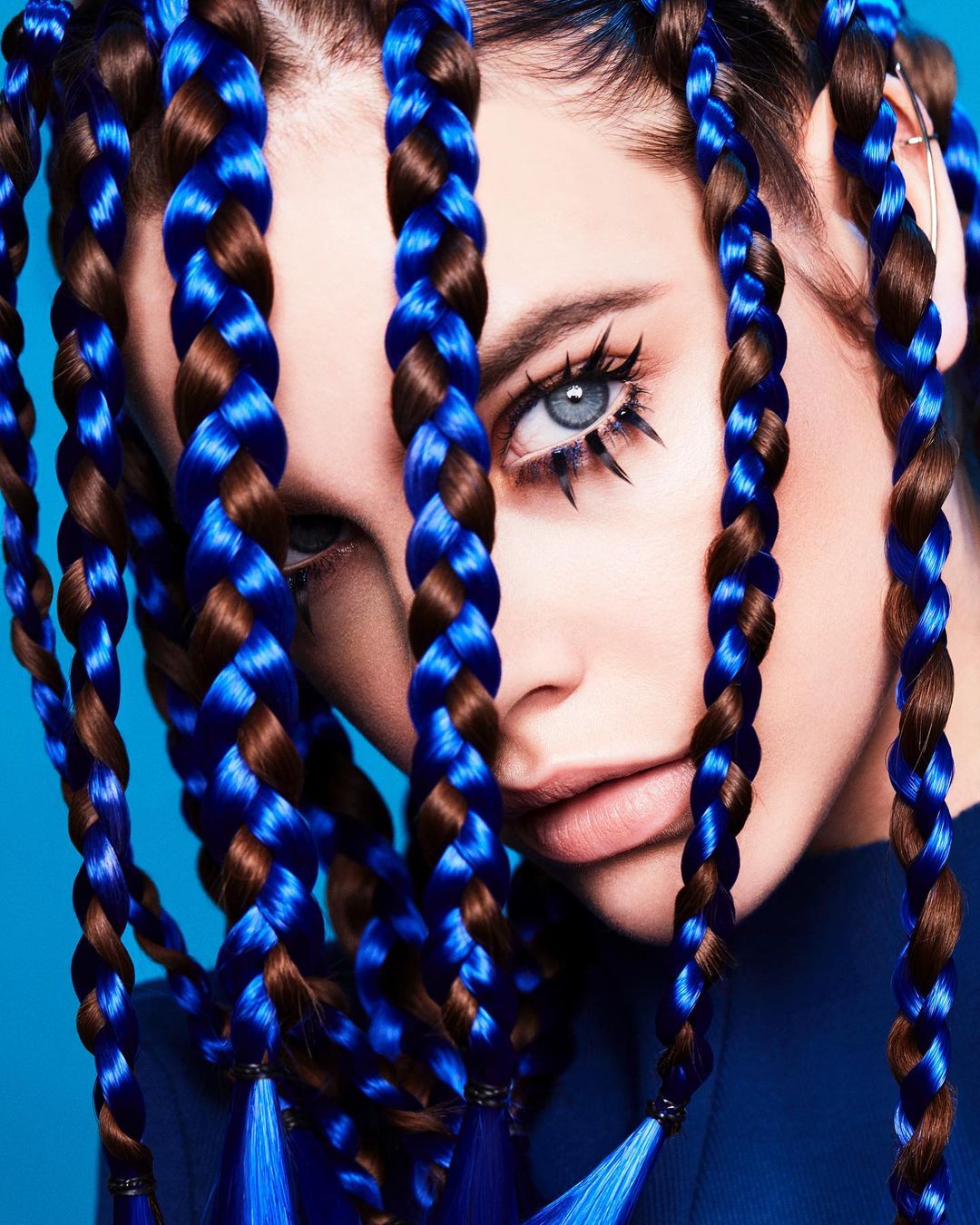 Portrait of a woman with blue eyes and brown hair styled in blue-and-brown braids, posing in front of a solid blue background.