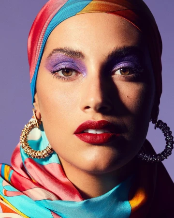 Close-up portrait of a woman with bright purple eyeshadow, red lipstick, and a colourful headscarf against a solid purple backdrop.