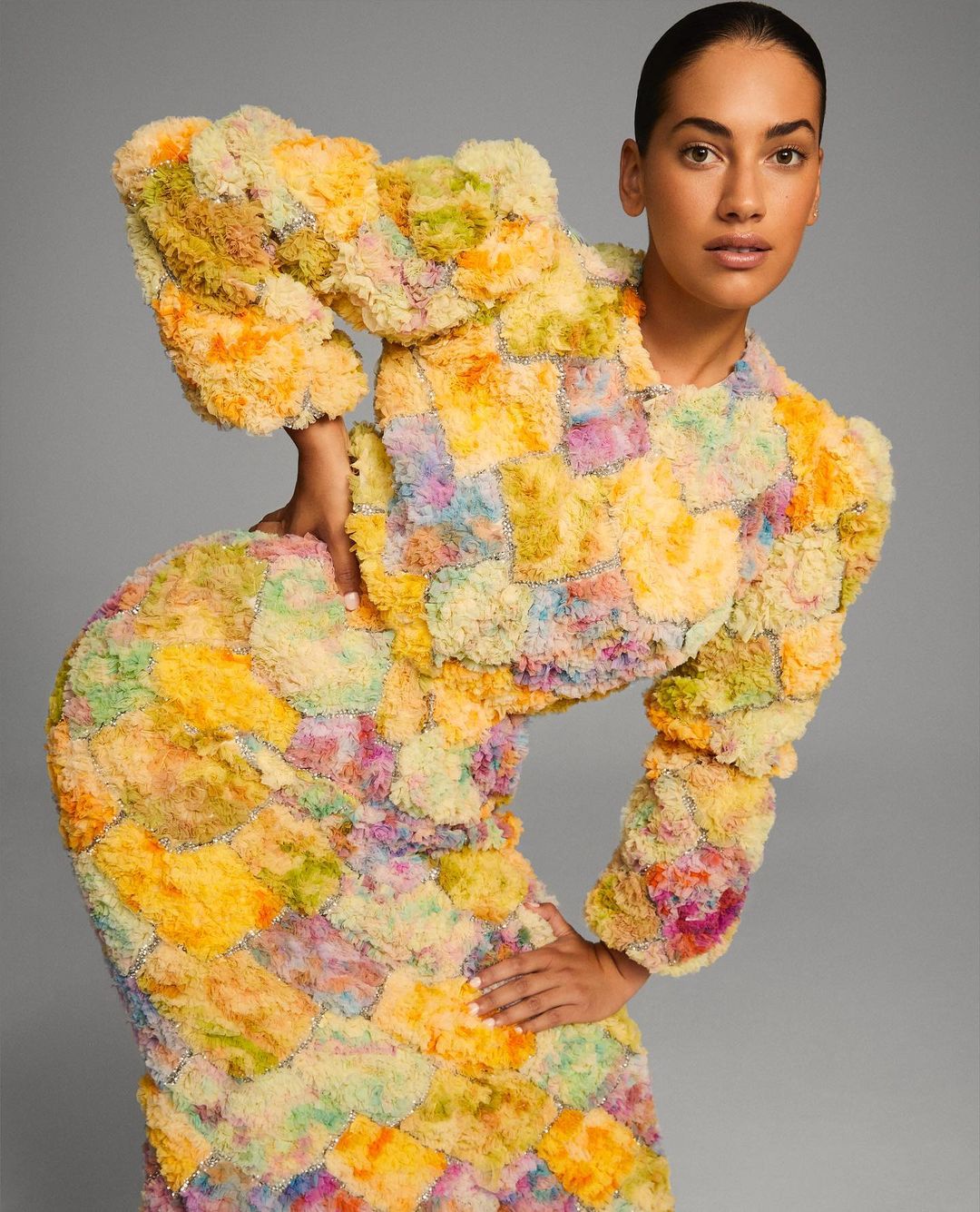 Woman posing in a colorful, textured patchwork dress in a studio. The dress features a short jacket with puffy sleeves and a fitted skirt.