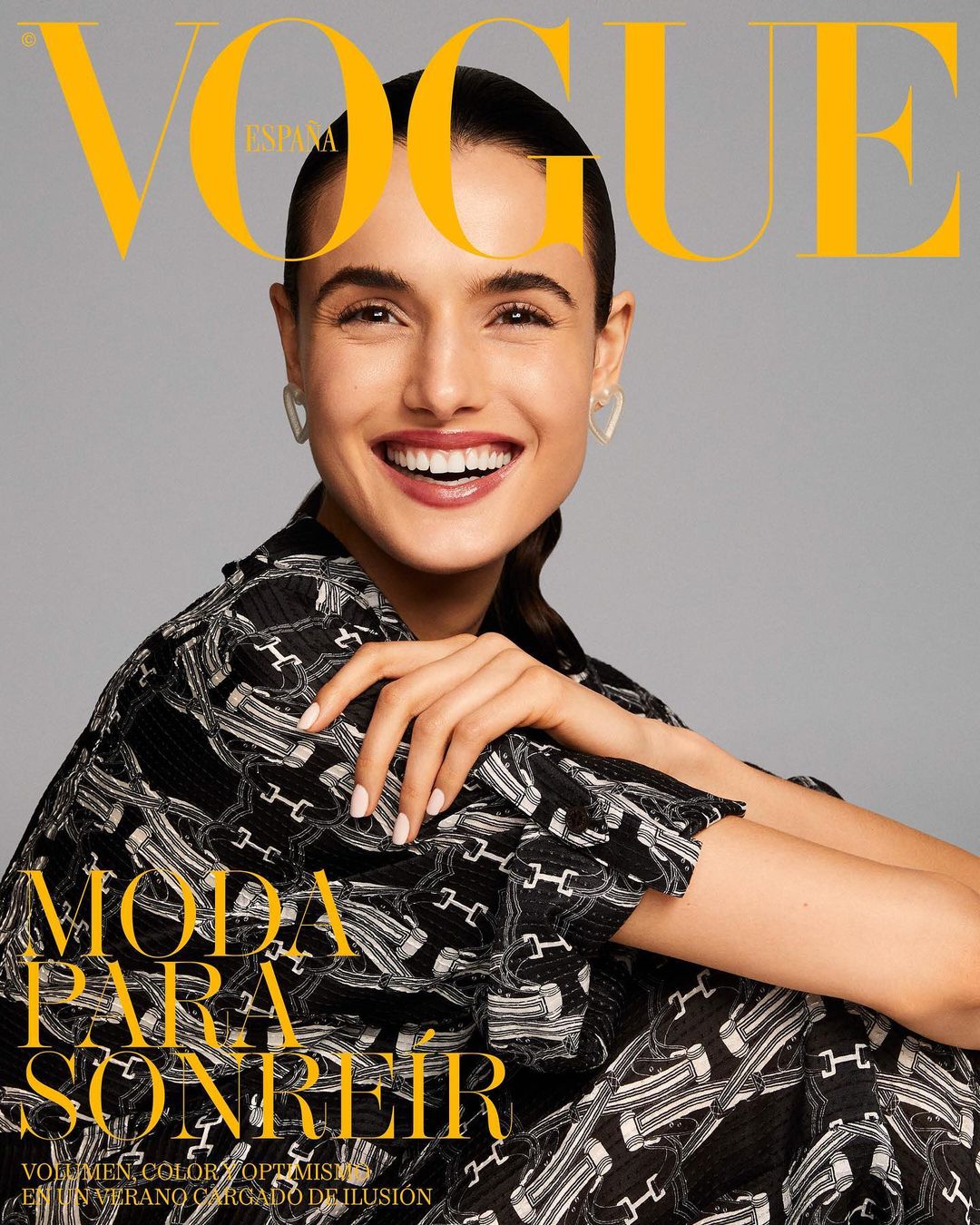 Vogue Espana cover featuring a smiling woman with heart earrings and a black patterned top against a gray background. Magazine cover.