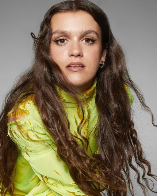 Close-up portrait of a young woman with long brown hair, wearing a light green floral top, against a light gray backdrop in a studio.