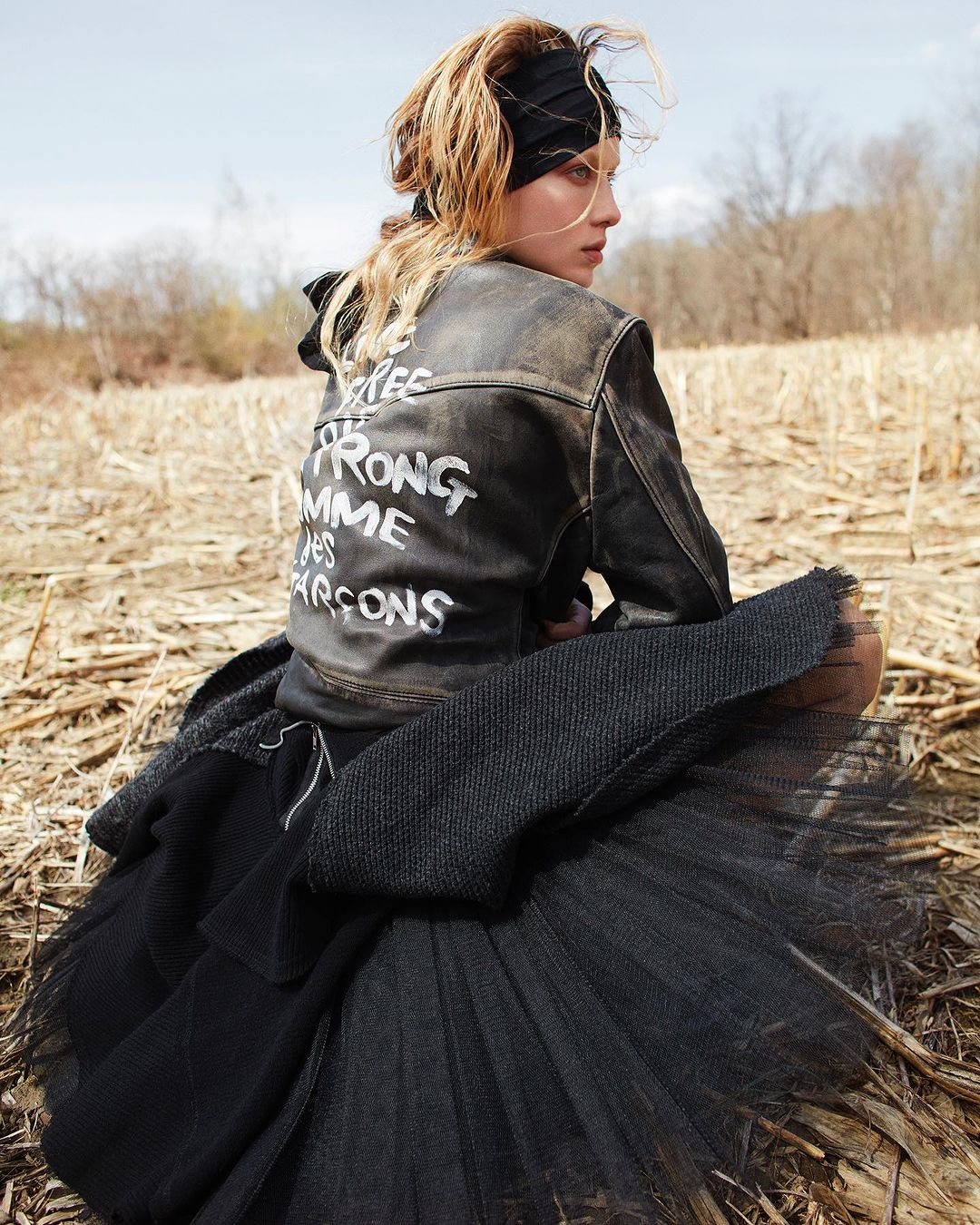 Woman with blonde hair in black tulle skirt and jacket, sitting in a field of dry grass in outdoor editorial fashion shoot.