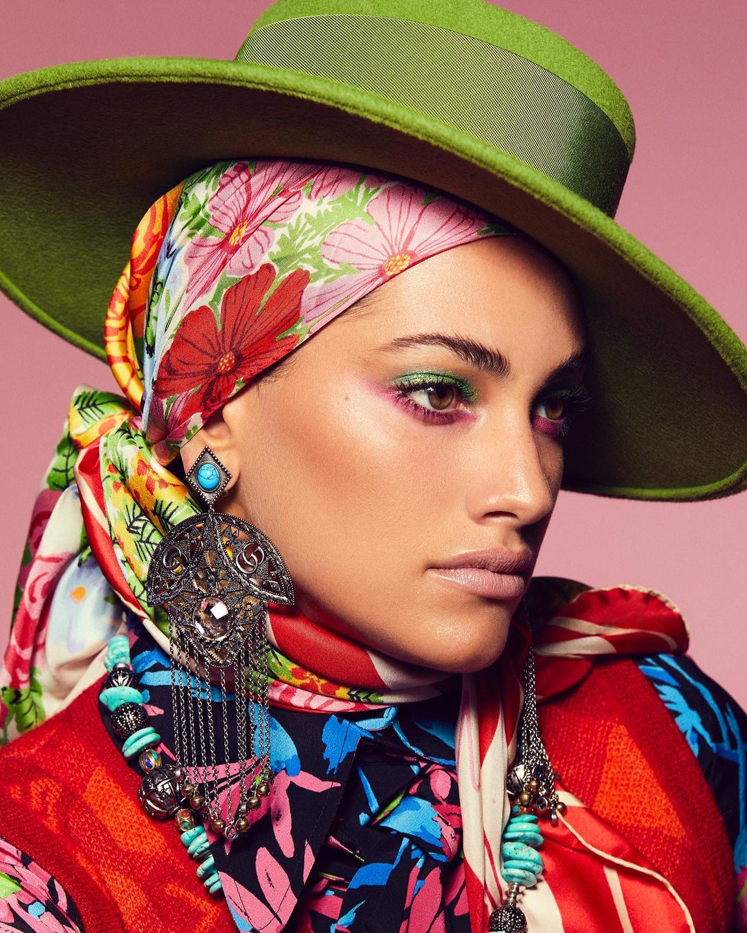 Portrait of a woman with a green hat, floral headscarf, and statement jewelry, showcasing vibrant makeup and a colorful outfit against pink.
