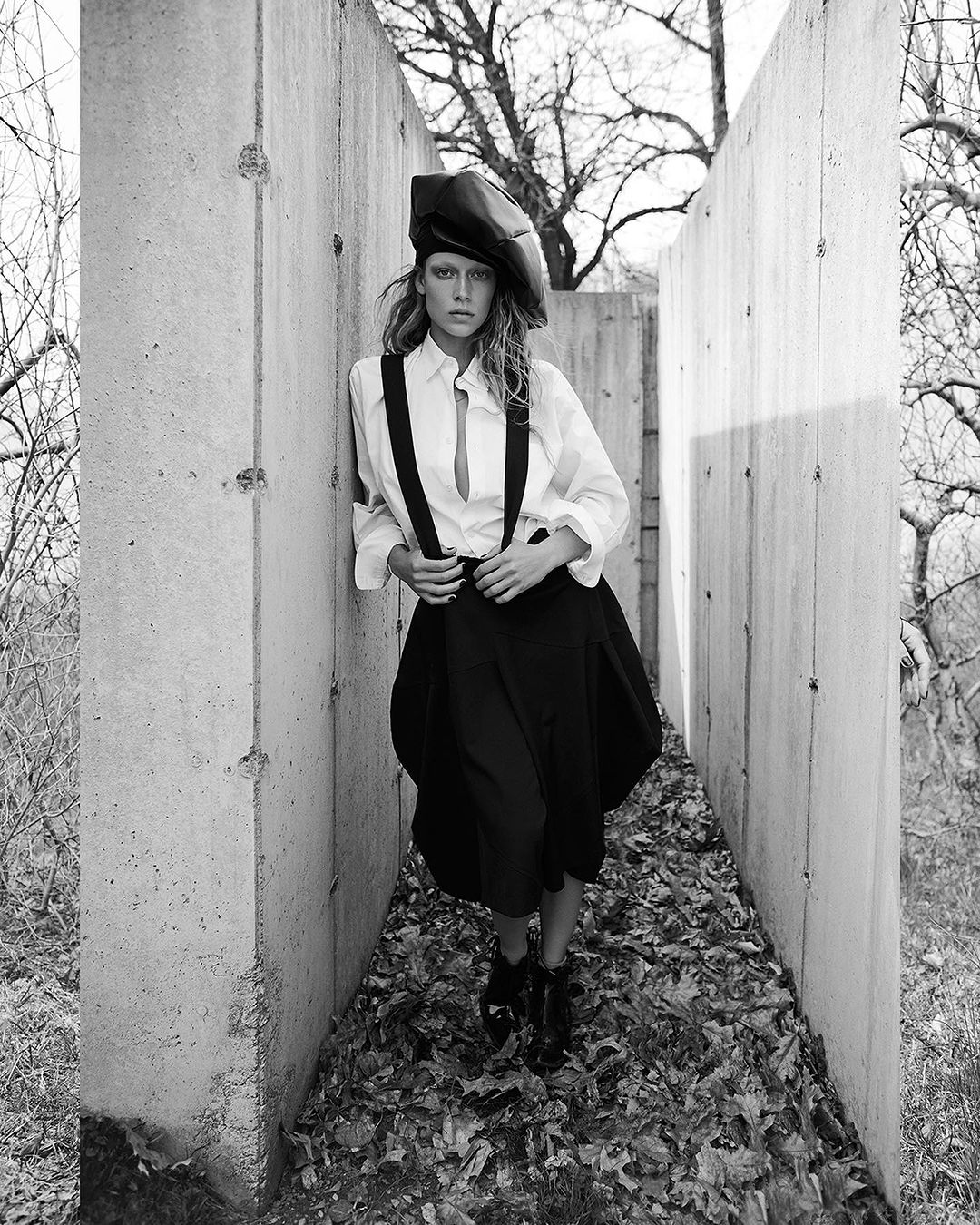 A woman in a black hat, suspenders, and skirt stands among concrete barriers and fallen leaves in a black-and-white fashion portrait.