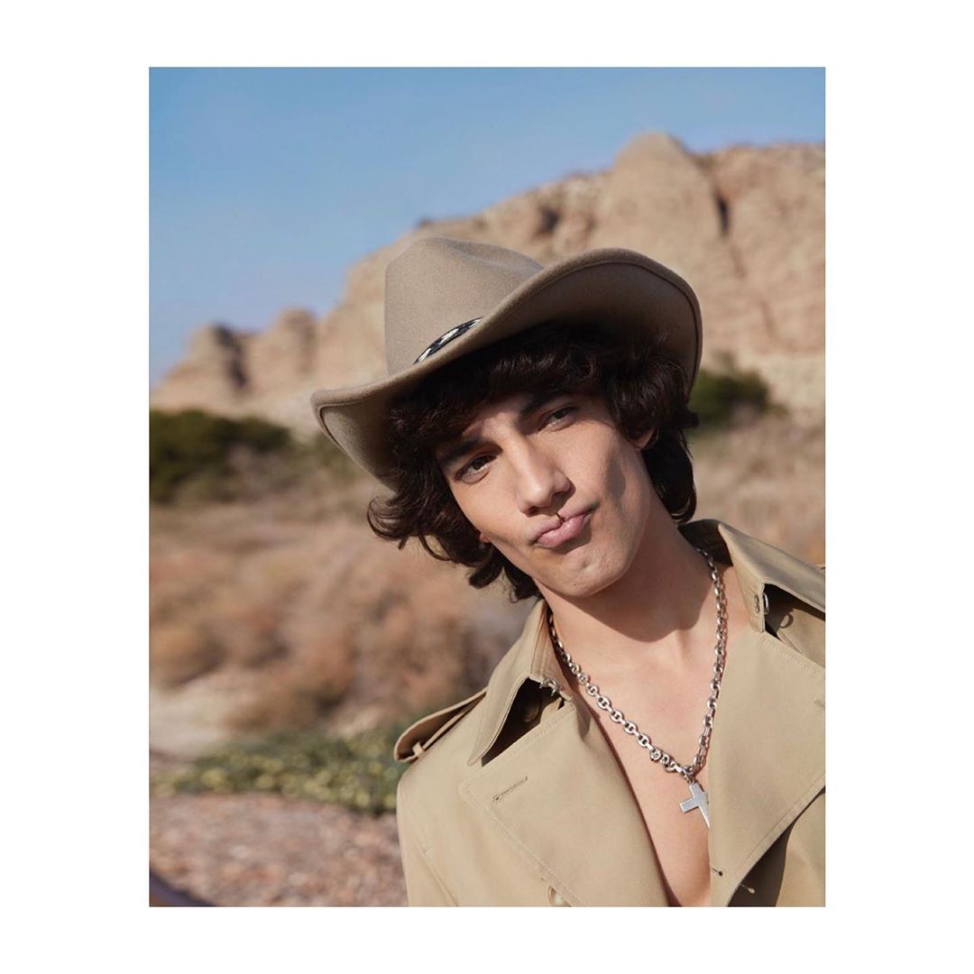 Close-up of a person in a cowboy hat and tan trench coat making a kissing face against a desert landscape with a blue sky.