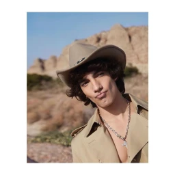 A young man wearing a cowboy hat and a tan coat with a silver cross necklace, set against an outdoor desert background.
