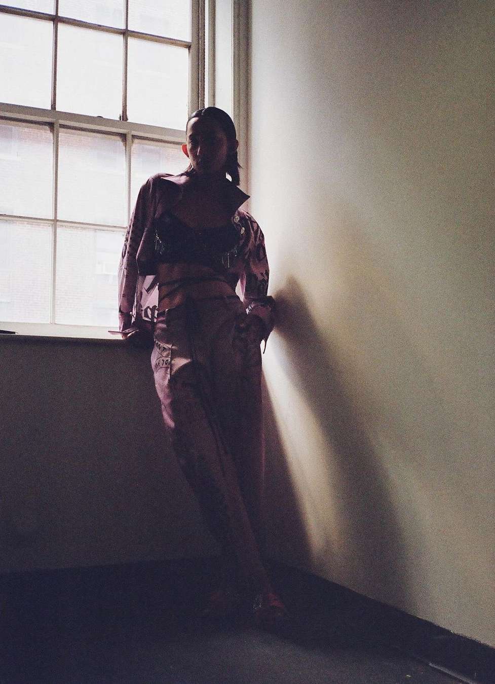 Woman in pink patterned outfit poses against window in dim light, blending fashion and architectural elements in a unique setting.