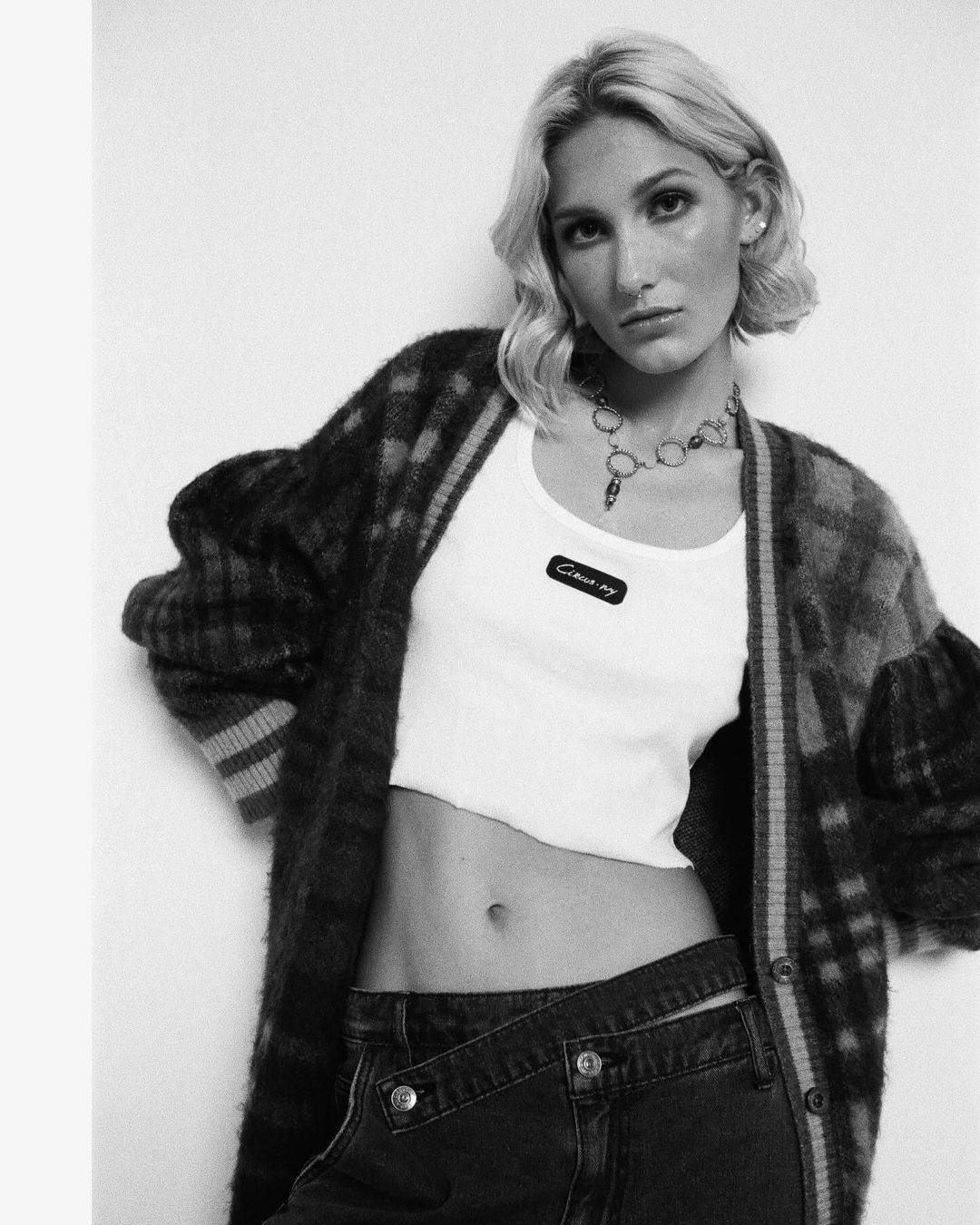 Black and white portrait of an adult woman with blonde hair wearing a cropped shirt, cardigan, and dark denim bottoms in studio.