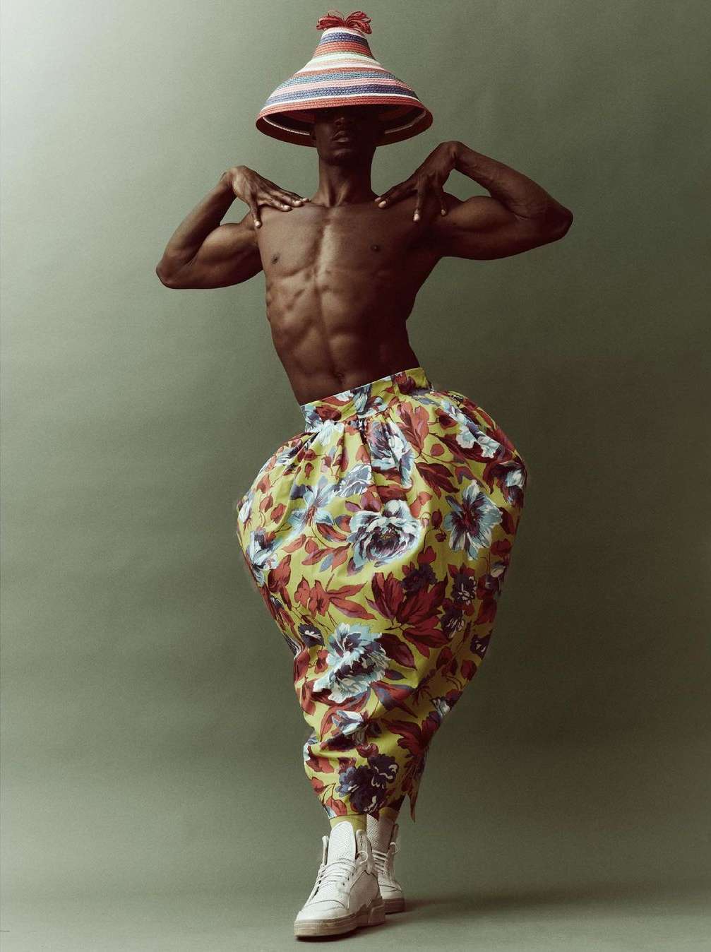 Shirtless man in floral pants and conical hat poses in studio, showing muscular physique and unique fashion style against green backdrop.