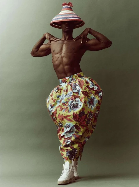 Shirtless man in floral pants and conical hat poses in studio, showing muscular physique and unique fashion style against green backdrop.