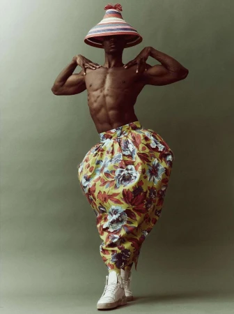 Shirtless man in floral pants and conical hat poses in studio, showing muscular physique and unique fashion style against green backdrop.