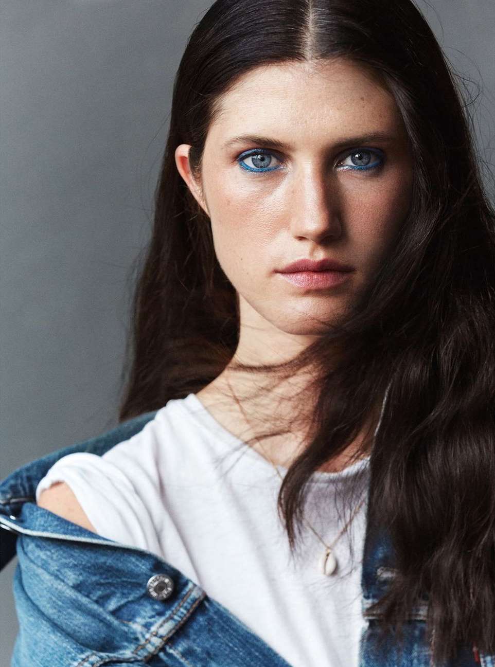 Portrait of a young adult woman with dark hair and blue eyeshadow, wearing a white t-shirt and a blue denim jacket. Grey background.