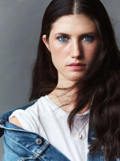 Portrait of a young adult woman with dark hair and blue eyeshadow, wearing a white t-shirt and a blue denim jacket. Grey background.