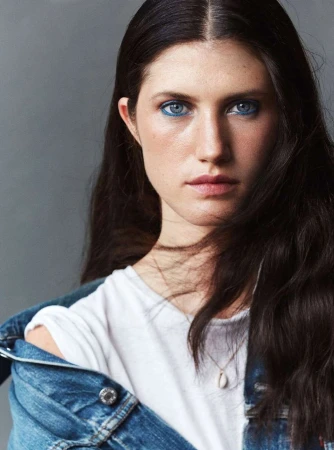 Close-up portrait of a woman with long dark hair, blue eye makeup, a white t-shirt, and a denim jacket draped over her shoulders.
