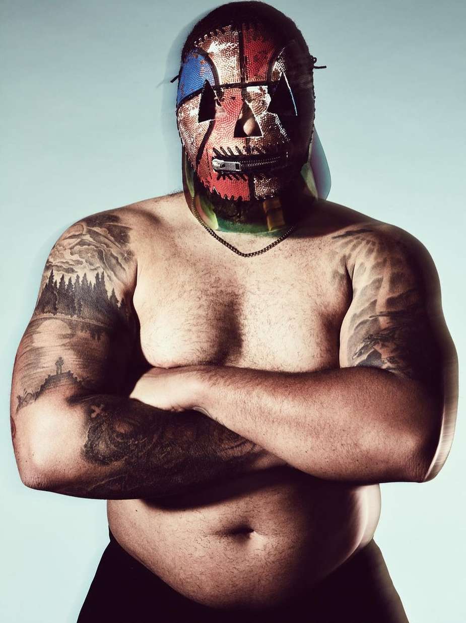 Man wearing a colorful stitched mask with a zipper mouth and heavy arm tattoos crossing his arms against a plain light background